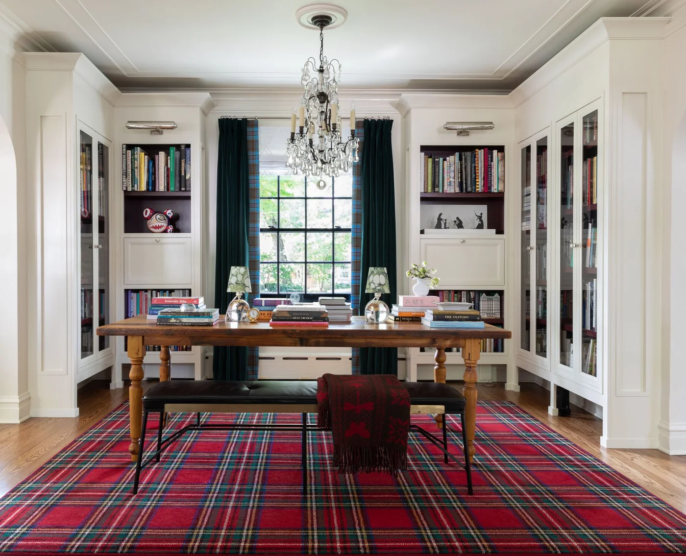 White glazed library cabinets and open shelves surround a long wooden table, dark bench and tartan rug near a tall window