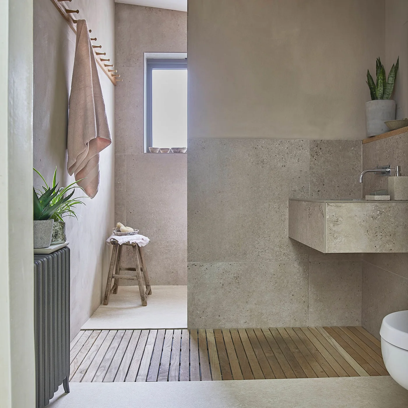 A pale textured wall basin and tiled lower walls sit beside muted plaster-like surfaces, wood floor slats, a small stool and hanging towels