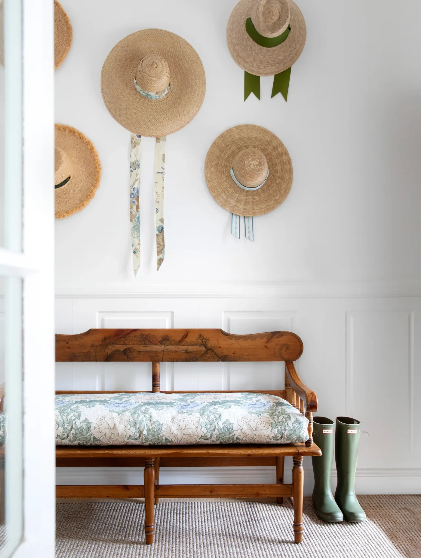 Country entry corner with a wooden bench, pale patterned seat cushion, woven hats on the white wall and boots beside the seat.
