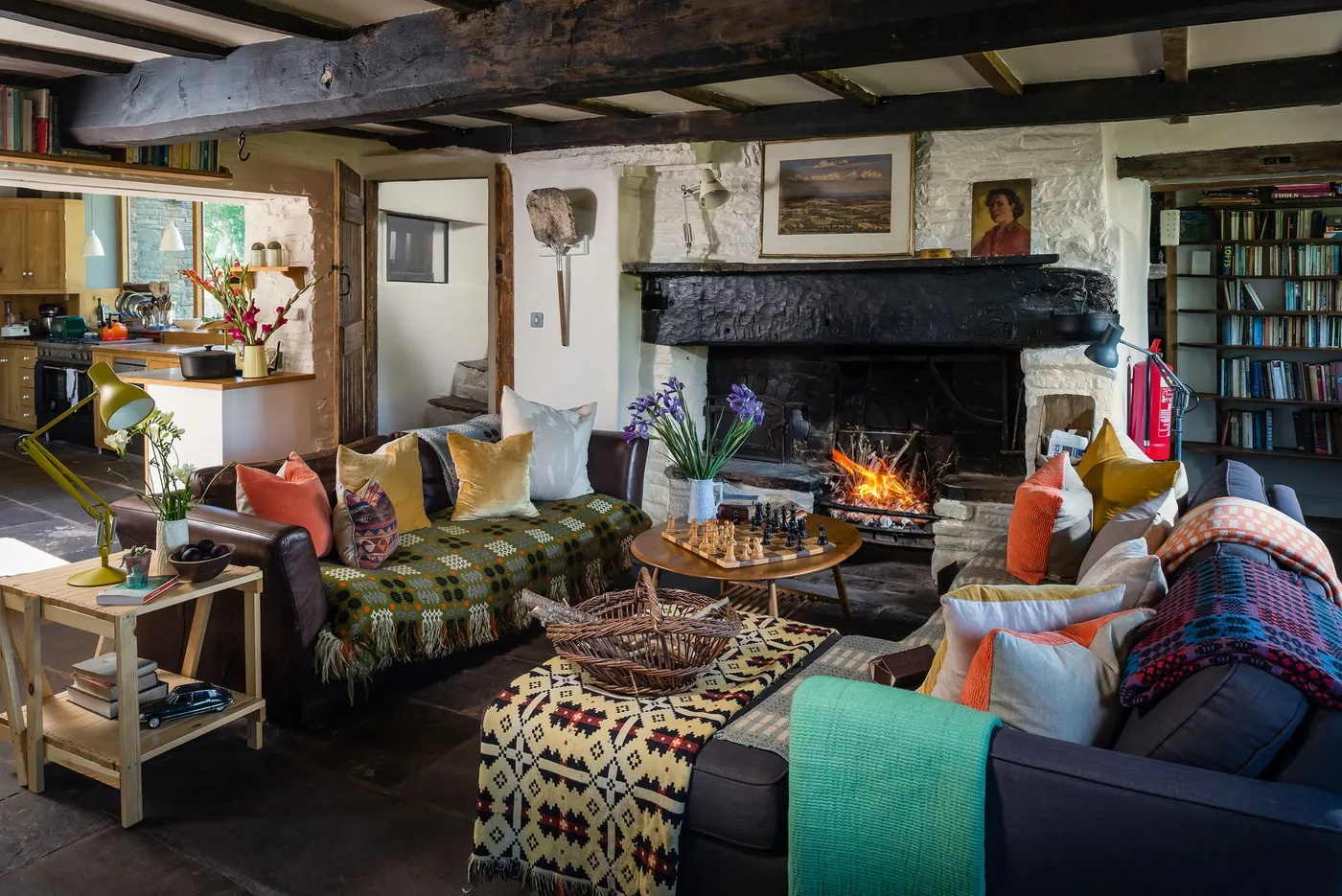 Broad dark fireplace beneath exposed ceiling beam with bookshelves, mixed seating and orange, yellow and patterned cushions.
