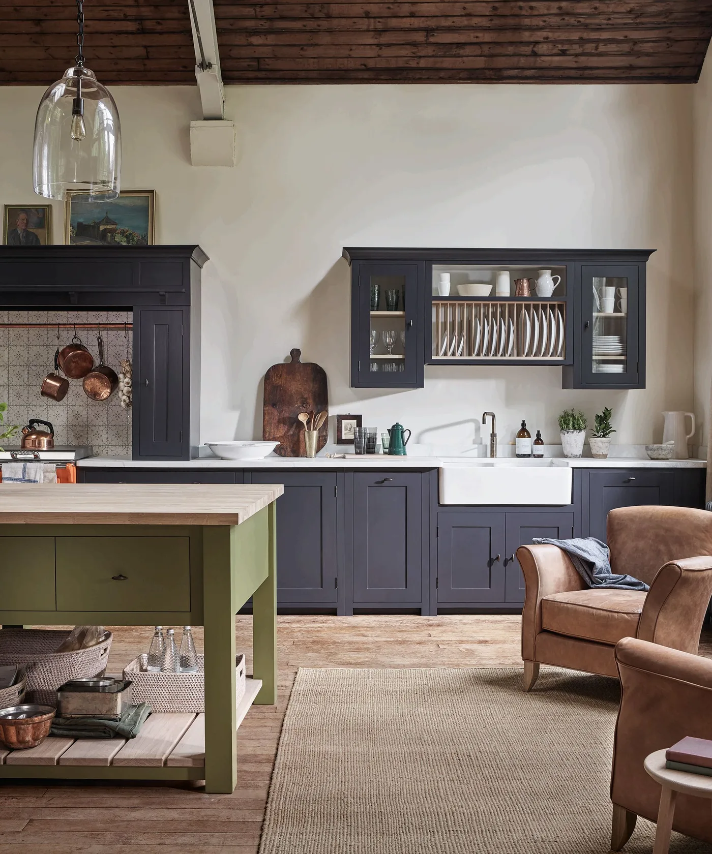 Kitchen with deep navy lower cupboards and glazed plate-rack wall cabinet, white sink, pale walls, wood ceiling, green island, and tan armchair.