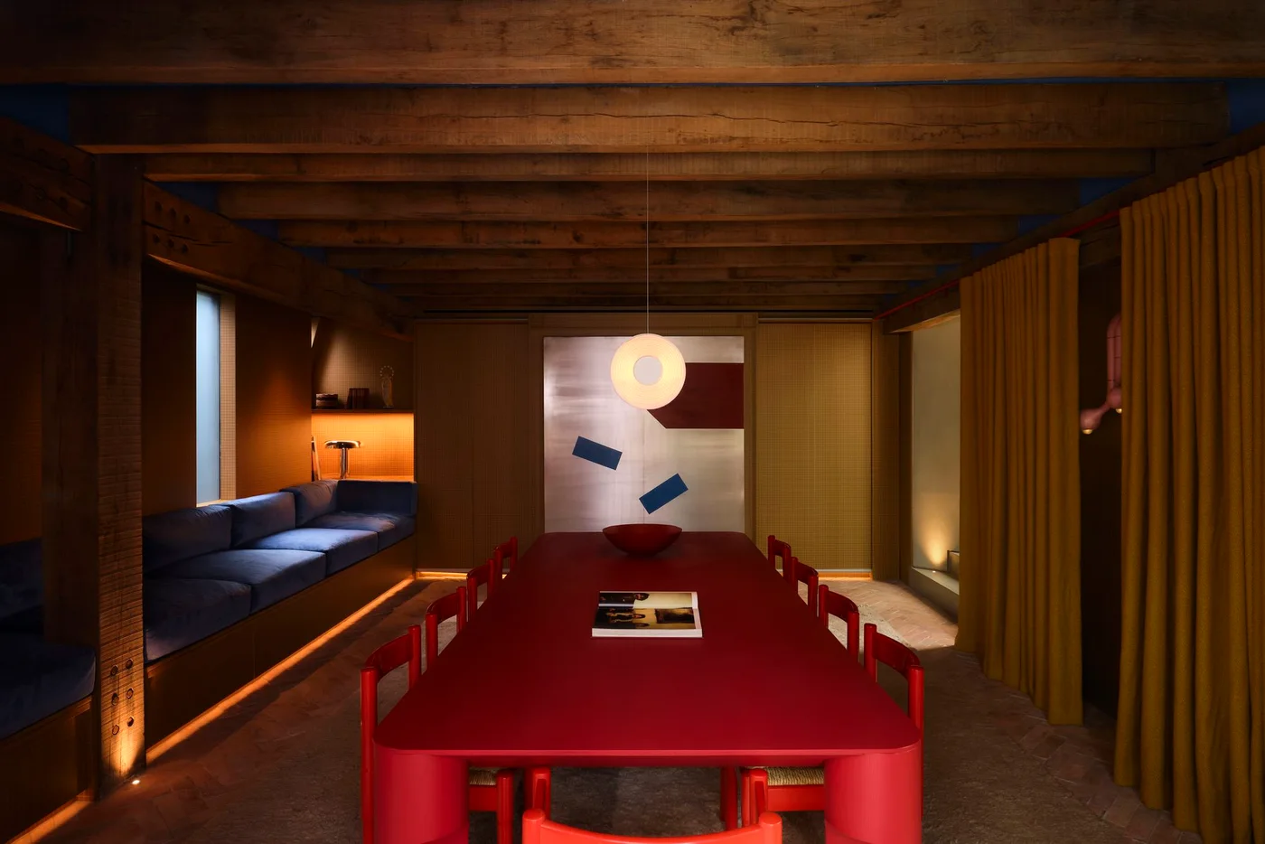 A red dining table and chairs sit under exposed wooden beams beside a long navy bench seat, warm walls and a yellow curtain