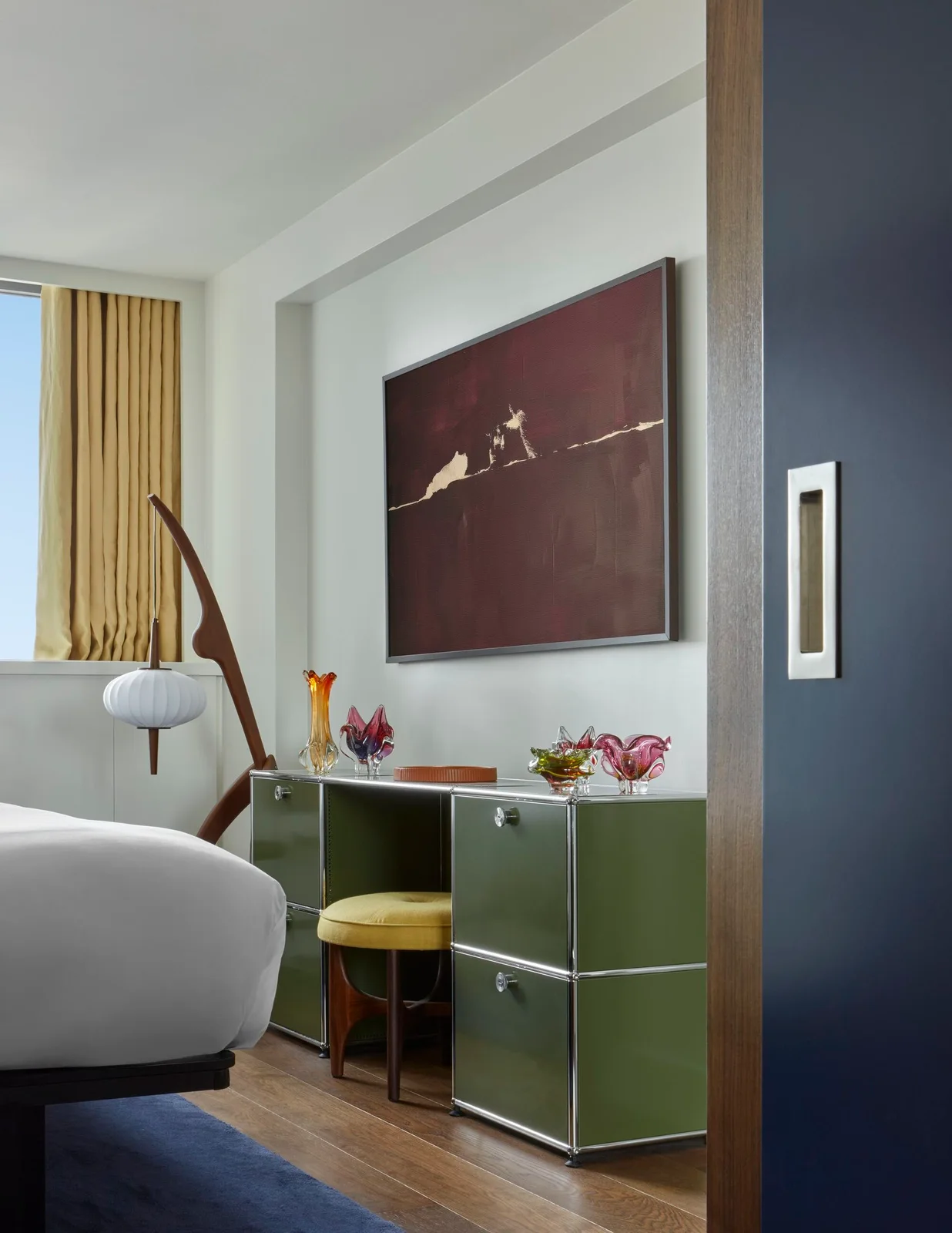 A green bedroom dressing table with drawers sits below framed artwork beside yellow curtains, a yellow seat and sculptural wood floor lamp