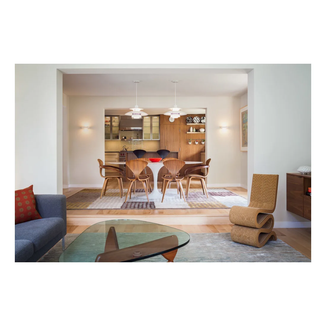 A wood dining table with curved wood chairs and two white pendant lights sits on a patterned rug beside kitchen cabinets and an adjoining sitting room