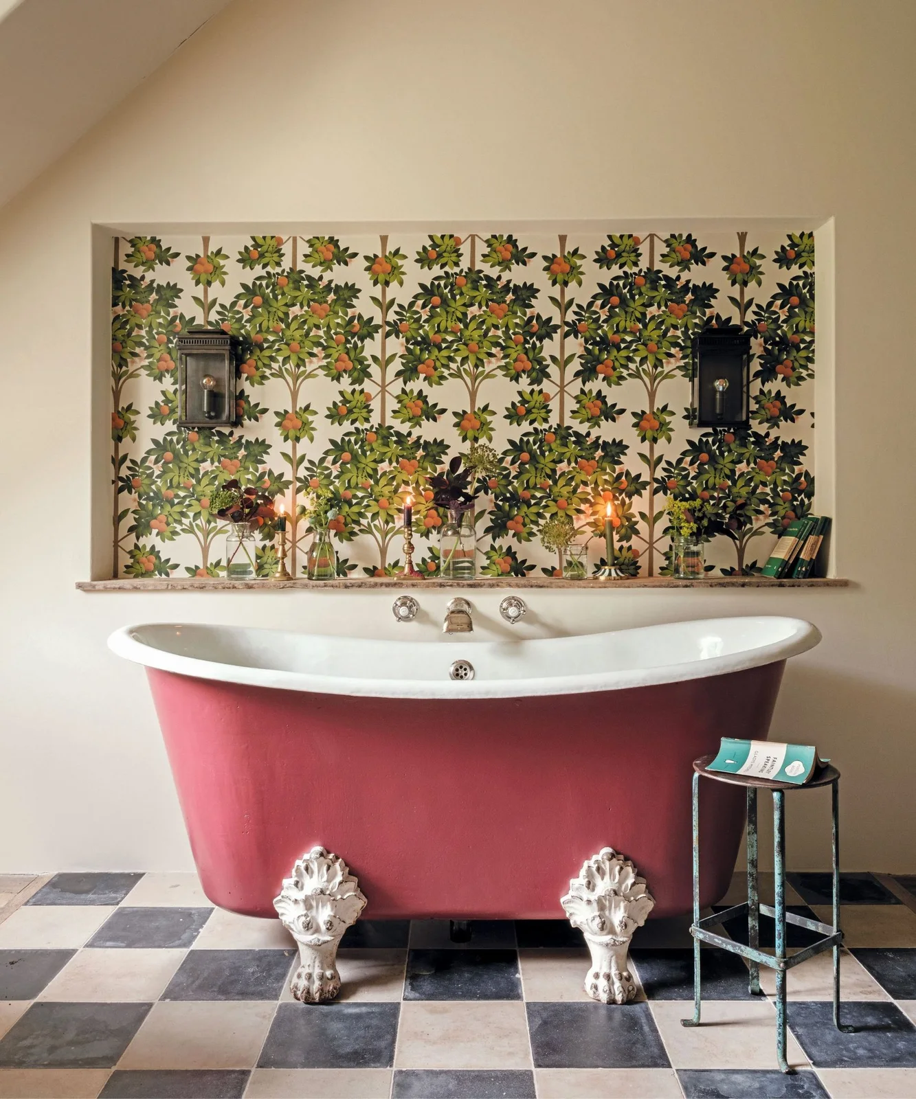A red claw-foot bath stands below botanical wallpaper and dark wall lights above a black-and-white checked floor