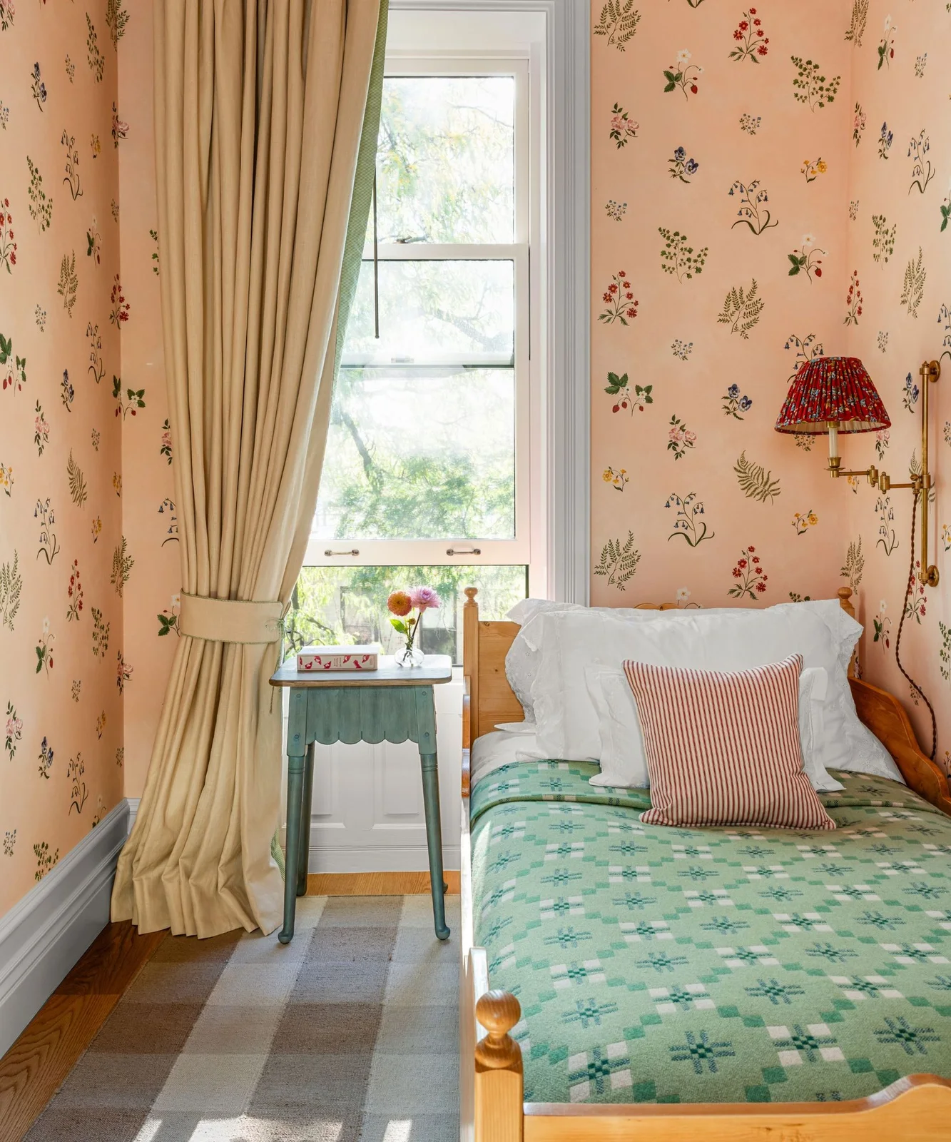 A single wooden bed sits against scattered floral wallpaper with a green checked blanket, striped pillow and patterned bedside wall-light shade