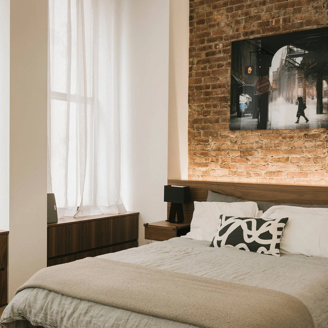 A wooden headboard and bedside cabinet sit against exposed brick beside white curtains, cream bedding and black-and-white artwork