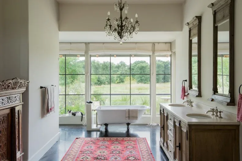 Long bathroom with dark wood double vanity, tall mirrors, red patterned runner, chandelier and clawfoot bath at broad windows