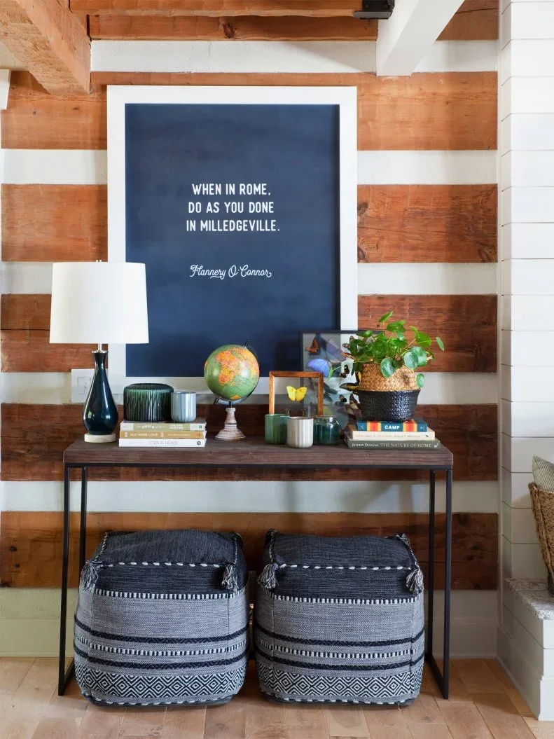 A slim dark entry console and two patterned storage baskets sit beneath alternating wood and pale wall boards, a large dark picture and a small shaded lamp