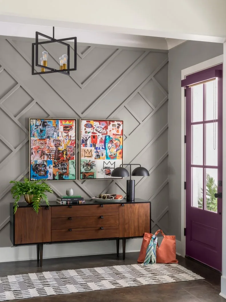 A warm wood entry cabinet with slim dark legs sits beside a gray geometric board wall, purple glazed door, dark lamps and a pale patterned rug
