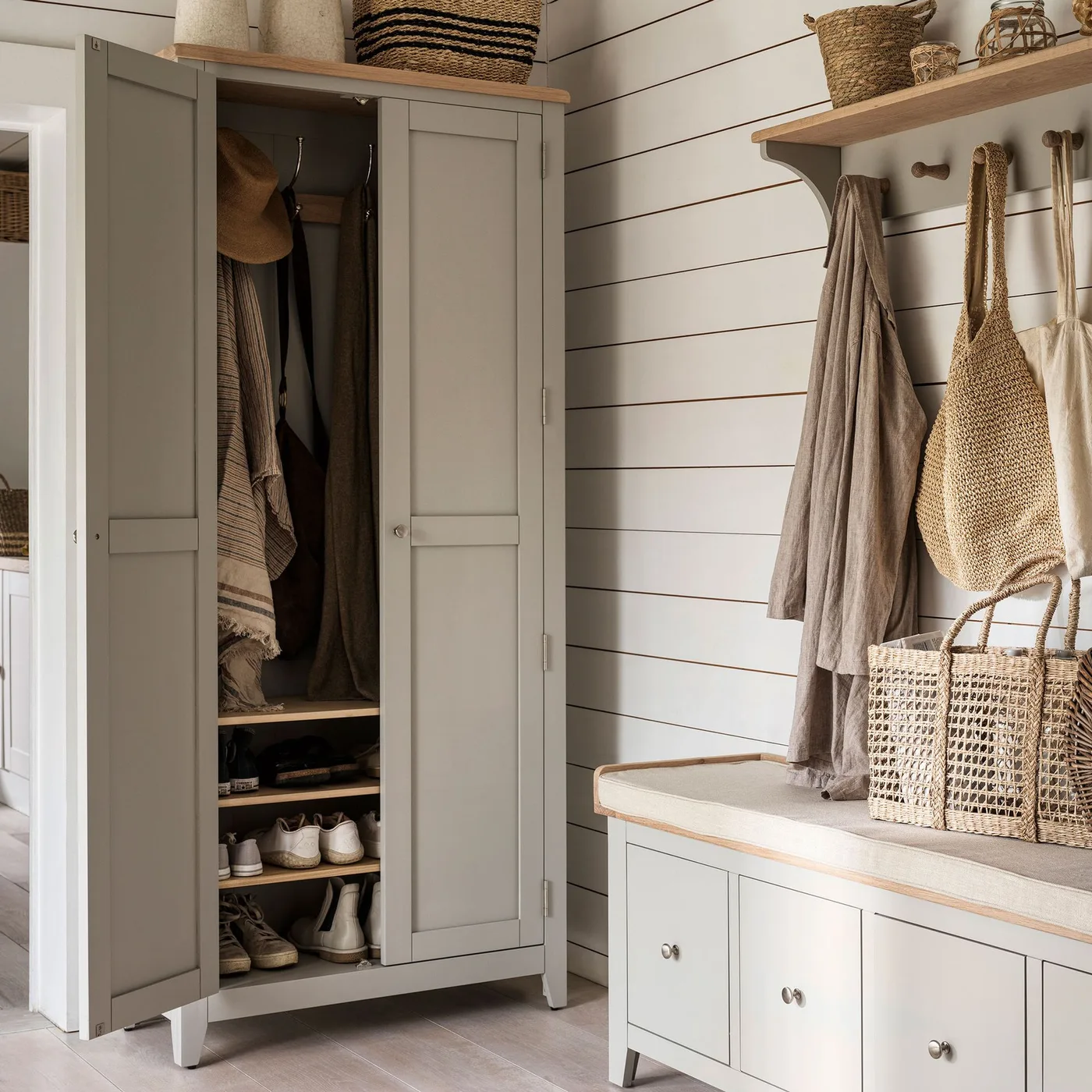 Country entrance with a gray coat cupboard, lower shoe shelves, matching drawer bench, wall pegs and upper wooden shelves holding baskets.