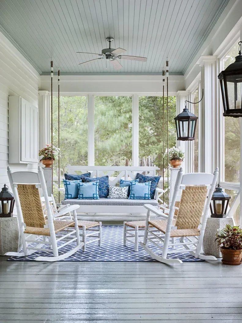 Screened white porch with suspended bench, blue cushions, woven-back rocking chairs and patterned rug