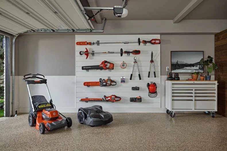 Garage slat wall with hanging yard tools beside a rolling cabinet and mower.