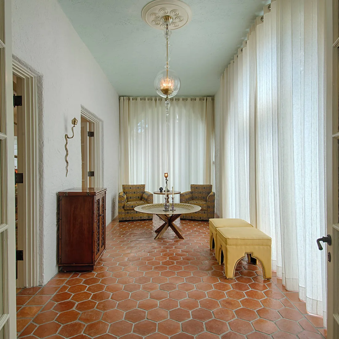 Warm hexagonal floor tiles lead toward two patterned chairs and a round table beneath a pale blue ceiling beside long light curtains and yellow stools