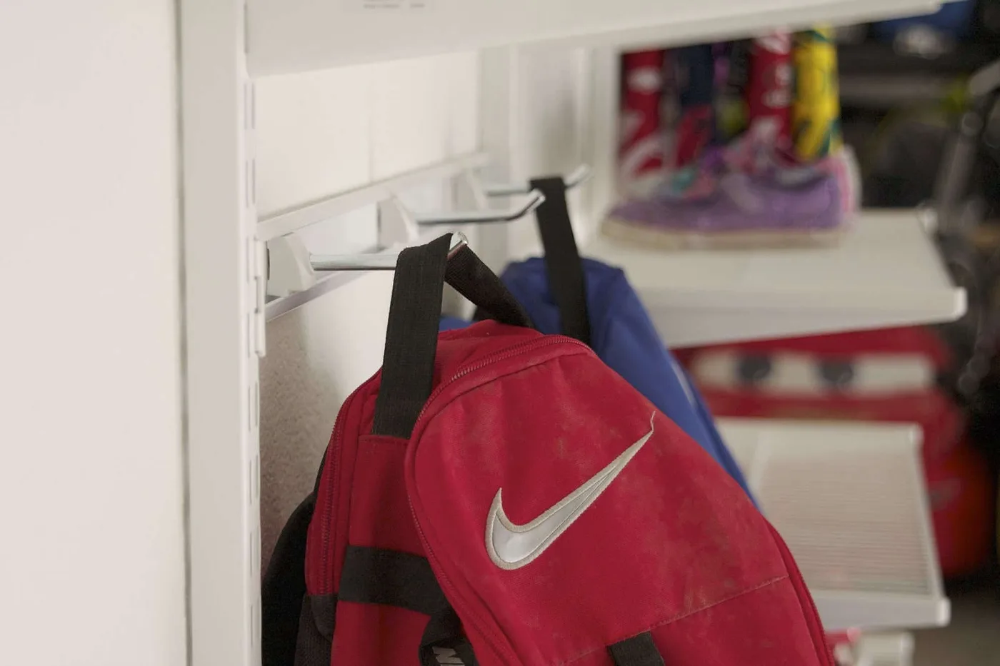 Red and blue backpacks hanging from white wall-mounted hooks beside open shoe shelving.
