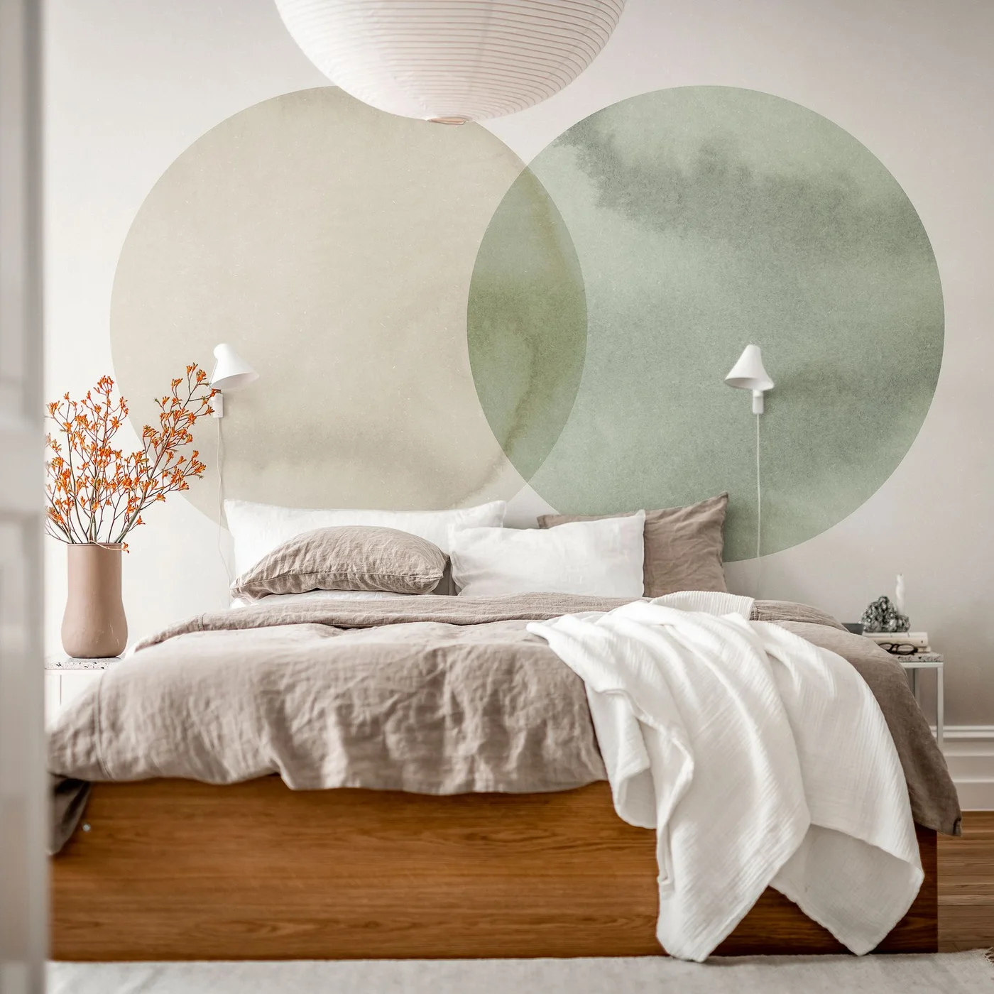 A low wooden bed with taupe and white bedding sits beneath overlapping muted green and beige wall circles and a large pale pendant