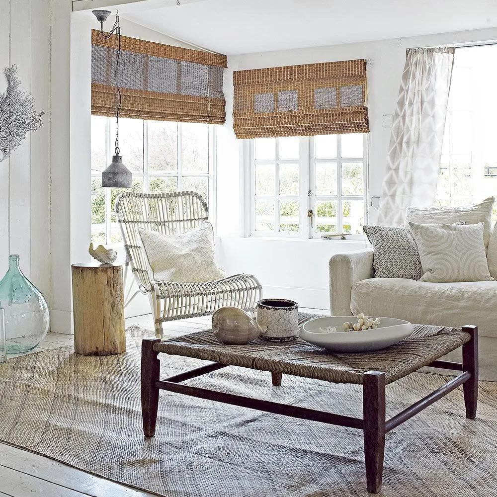 A pale sofa sits beside an open woven chair, a woven-top coffee table and warm woven window shades against light walls