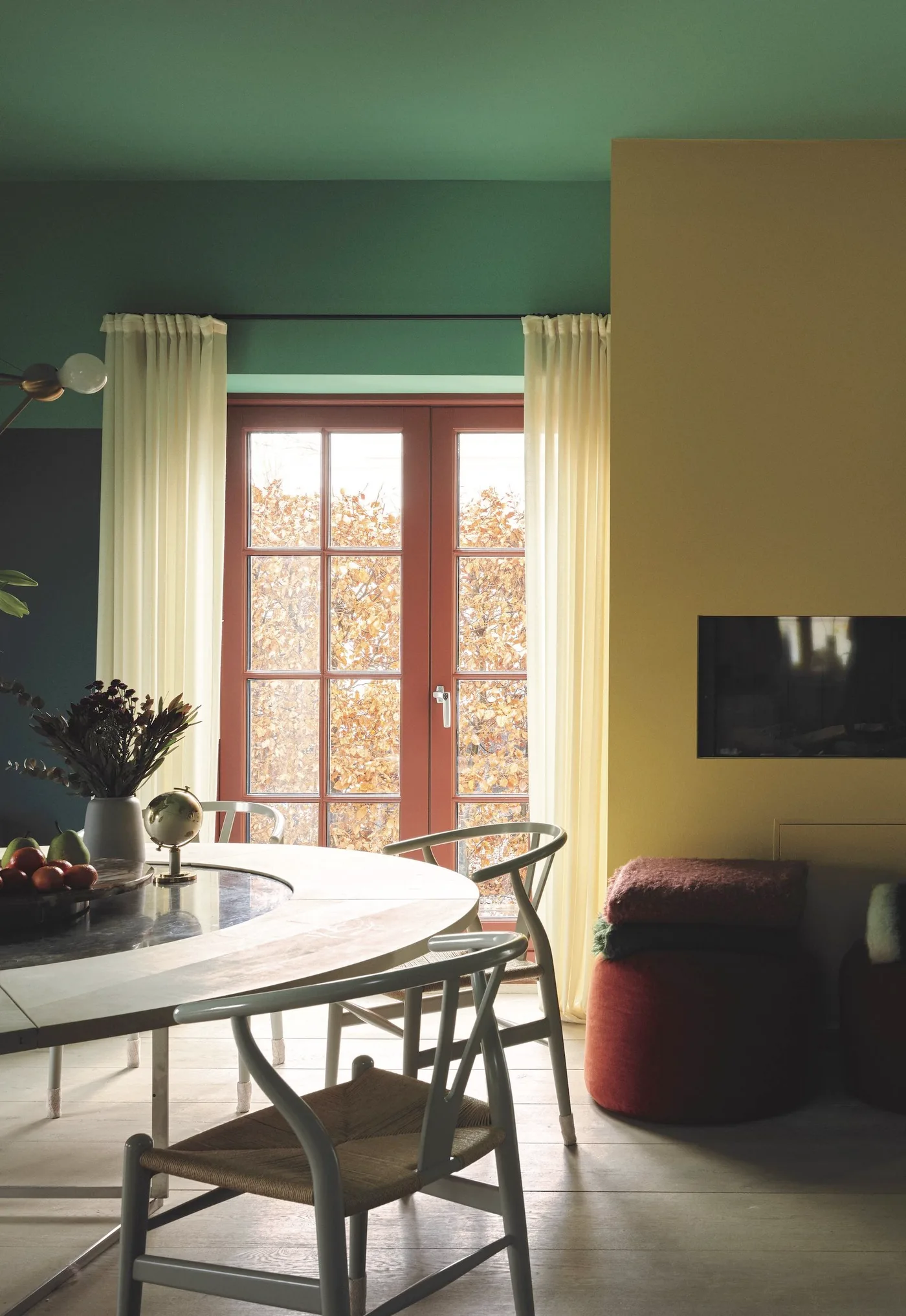Dining area with green walls and ceiling, a yellow wall, pale curtains, a round table and open-backed chairs