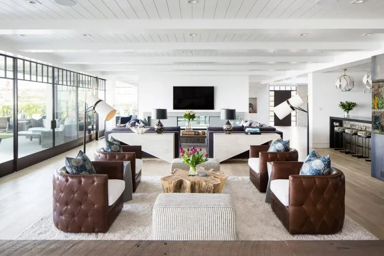 Large living room with brown low chairs and pale cushions around a wooden table, striped ottoman, pale sofas farther back and a broad wall of black-framed windows.