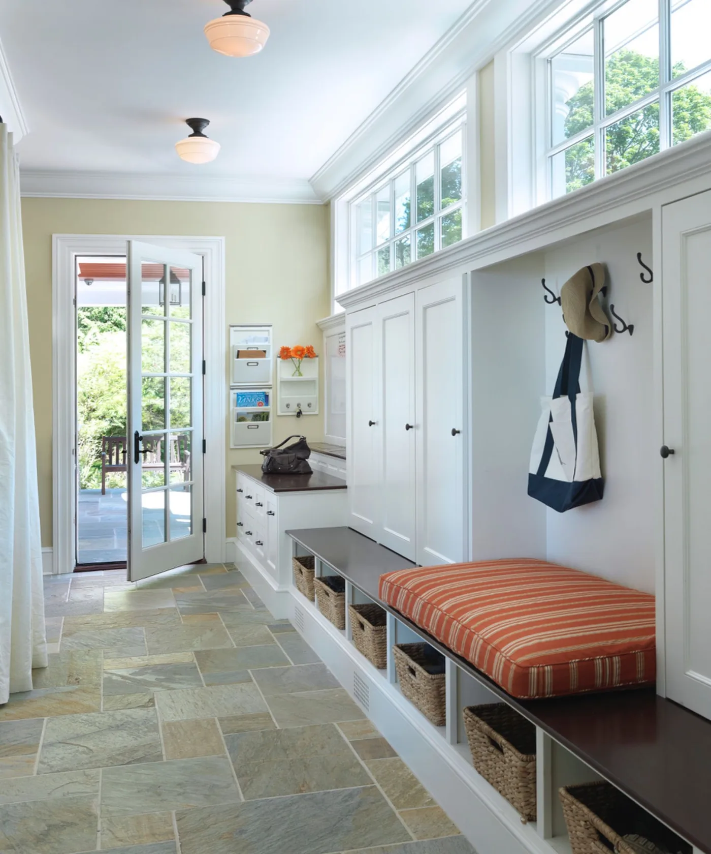 Long entry mudroom with high windows, white cupboards, wall hooks, wood bench, lower woven baskets, striped orange cushion and glazed door