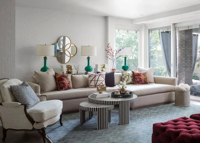 Living room with pale sofa and armchair, green lamp bases, patterned cushions, nested striped tables, blue-gray rug and red tufted ottoman edge.