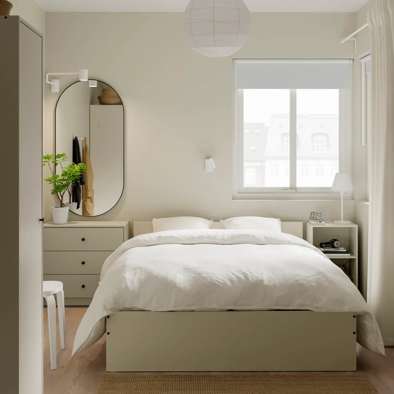 A plain pale bed sits beneath a window beside a drawer unit, oval mirror and small bedside shelf.