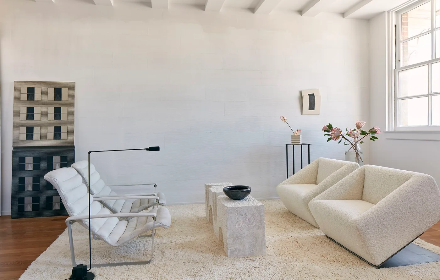 Two pale angular armchairs face two light-framed lounge chairs across a narrow pale table on a warm wood floor beneath plain light walls