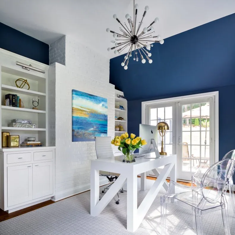 White desk and built-in shelving in a navy office with a starburst chandelier and clear guest chairs.