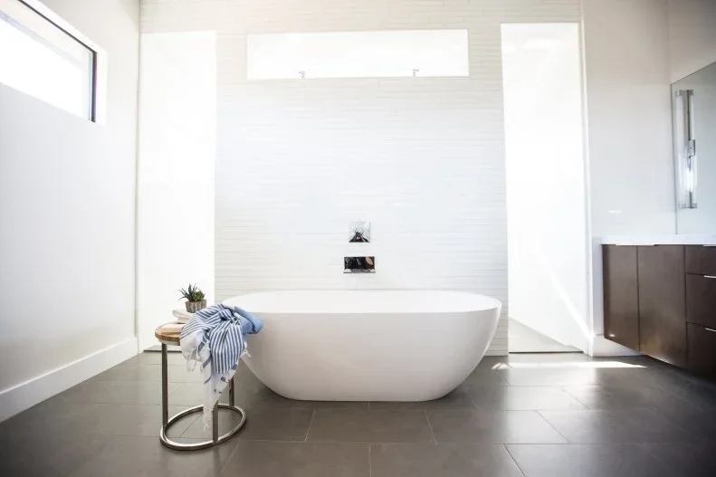 A white freestanding bathtub stands on a dark gray tiled floor below white horizontal wall tiles and a high window beside a narrow metal stand