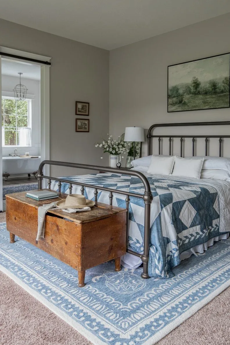 Gray bedroom with dark metal bed, blue patterned quilt, wood chest and blue rug