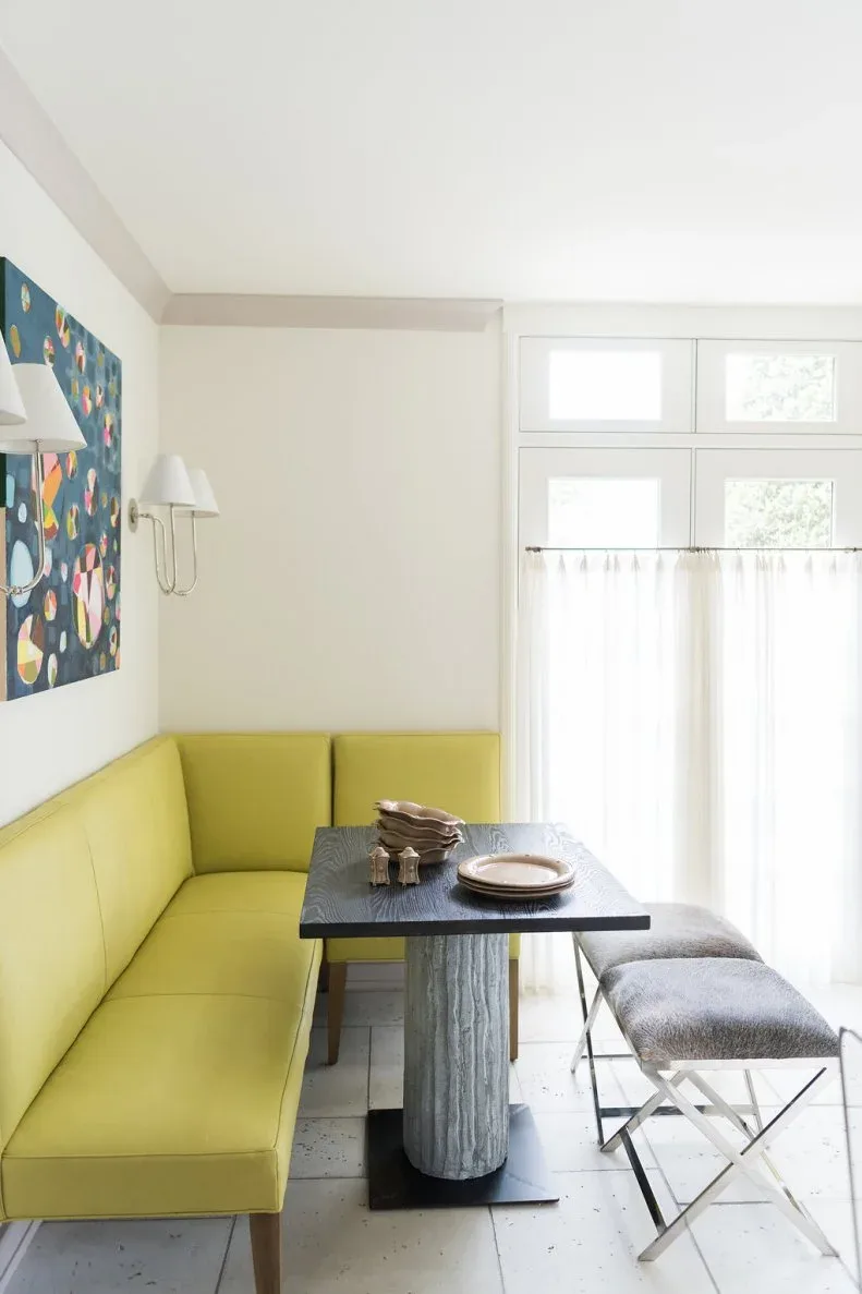 Yellow upholstered corner banquette beside a dark rectangular pedestal table, with gray stools on metal cross-shaped legs, pale walls, artwork, and a curtained window.