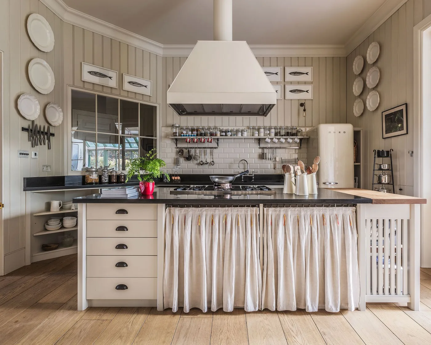 Cottage kitchen with pale vertical paneling, light drawers, a gathered white island skirt, dark countertop and a large pale hood above the cooking area.
