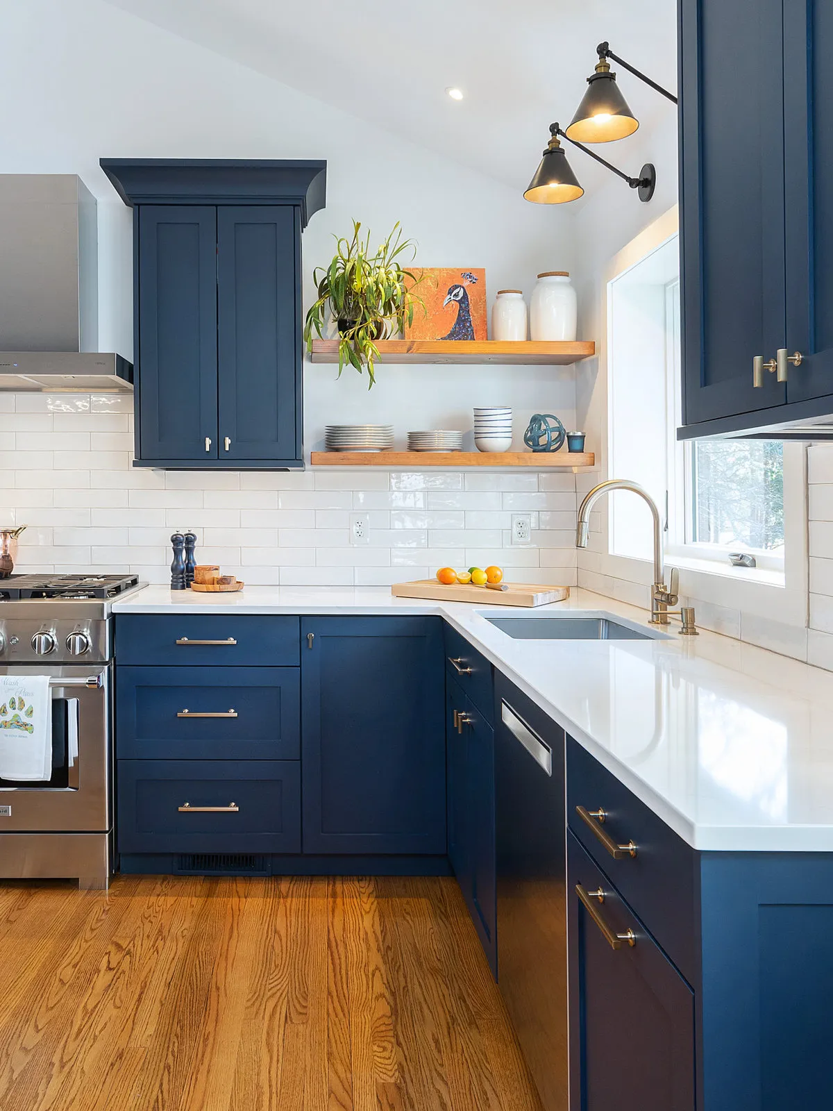 Navy upper and lower cabinets surround wooden open shelves of dishes beside a sink, white wall tiles and adjustable dark wall lamps