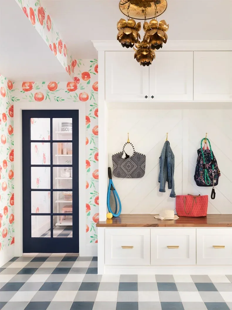 Mudroom with floral walls, white cabinets and bench drawers, dark glazed door, checked floor and gold-colored hanging light.