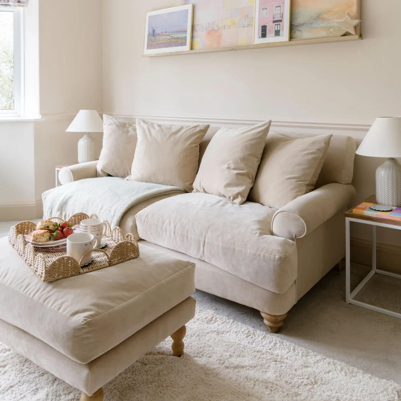 A pale sofa and matching footstool sit beneath a narrow picture ledge, with simple side lamps and a woven tray on the upholstered top