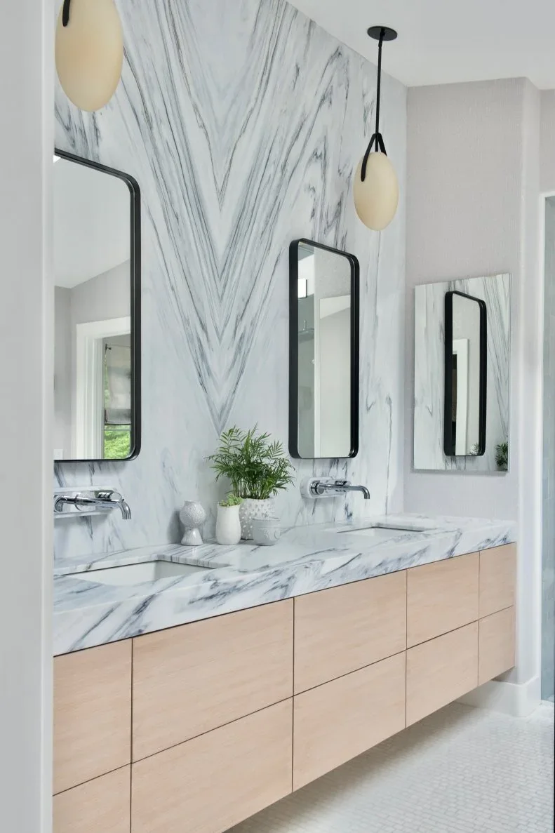 A long floating pale wood vanity stands below separate dark-framed mirrors and small hanging lights against a tall pale veined wall