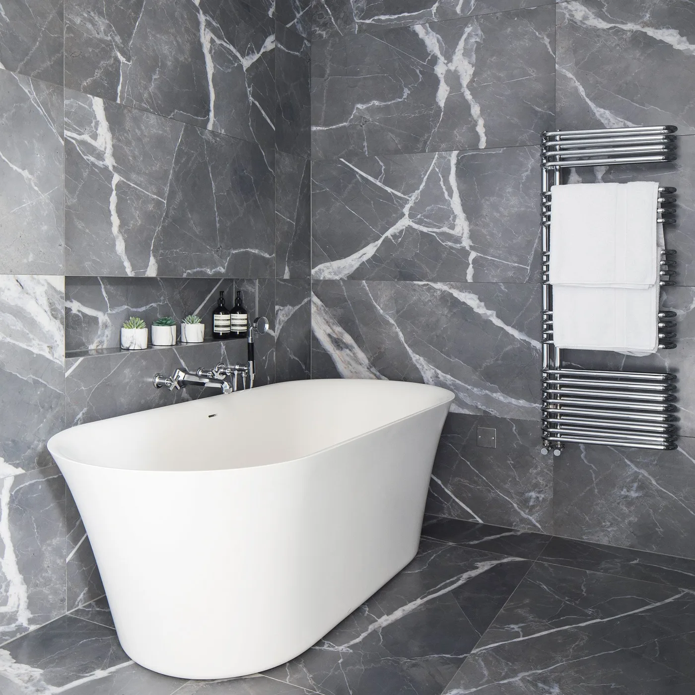 Bathroom with large deep gray white-veined wall and floor surfaces, white oval tub, reflective towel radiator, and recessed toiletries niche.