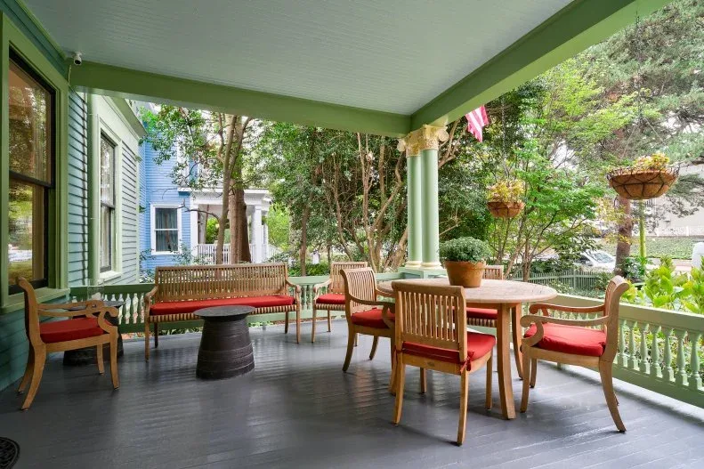 Green porch with wood seating, red cushions, round dining table and hanging baskets
