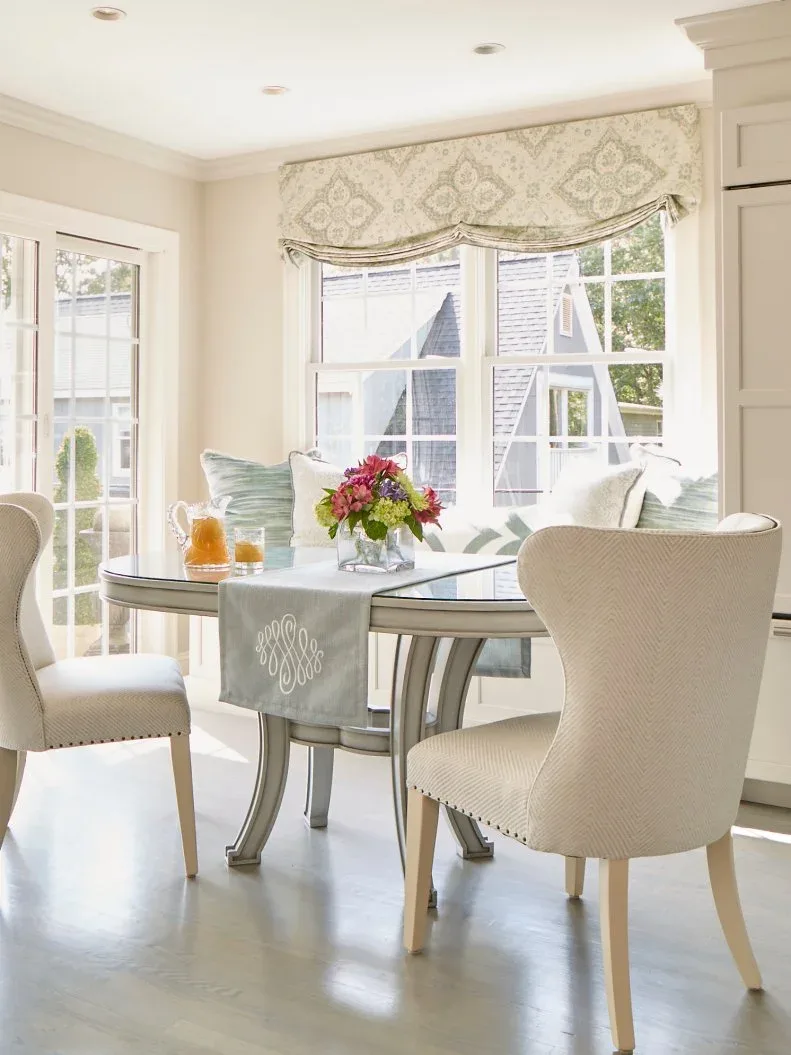 Pale rounded upholstered dining chair with nailhead edging beside a glass-topped table, another pale chair, window bench, and bright cottage windows.