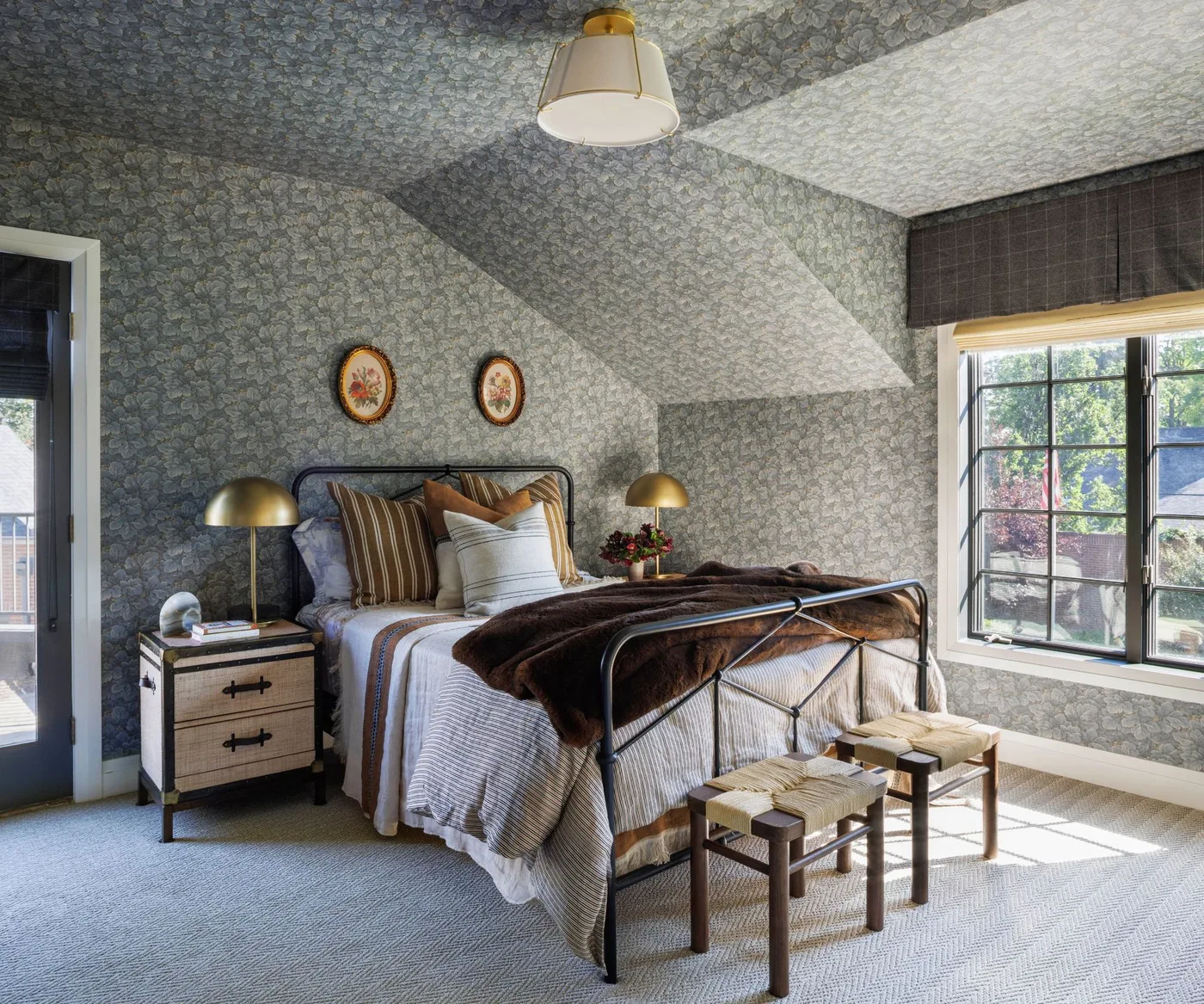 Gray floral wallpaper across bedroom walls and ceiling above a dark metal bed and two small stools.