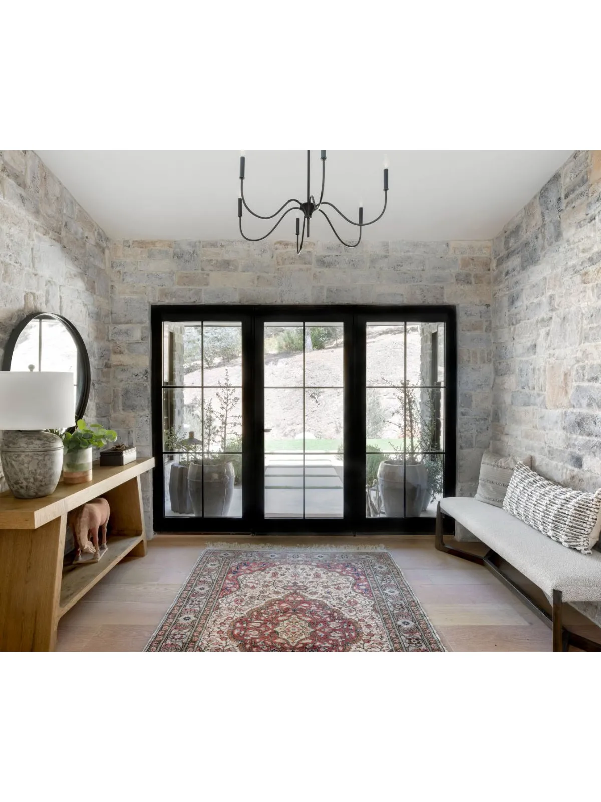 Patterned rug between a timber console and pale bench near dark-framed doors and textured stone walls