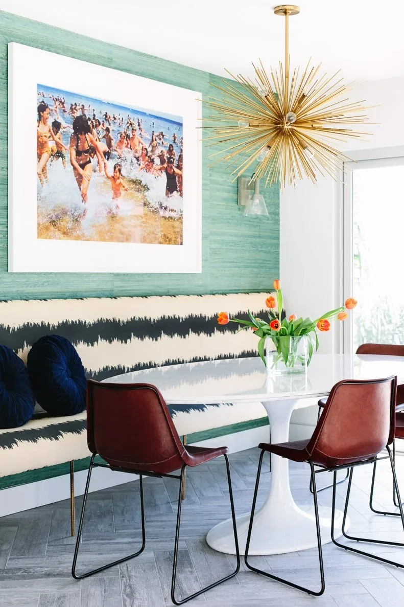 Black-and-white striped banquette beneath framed beach imagery on a green wall, with a white pedestal table, red-brown chairs, and a gold-colored starburst light.