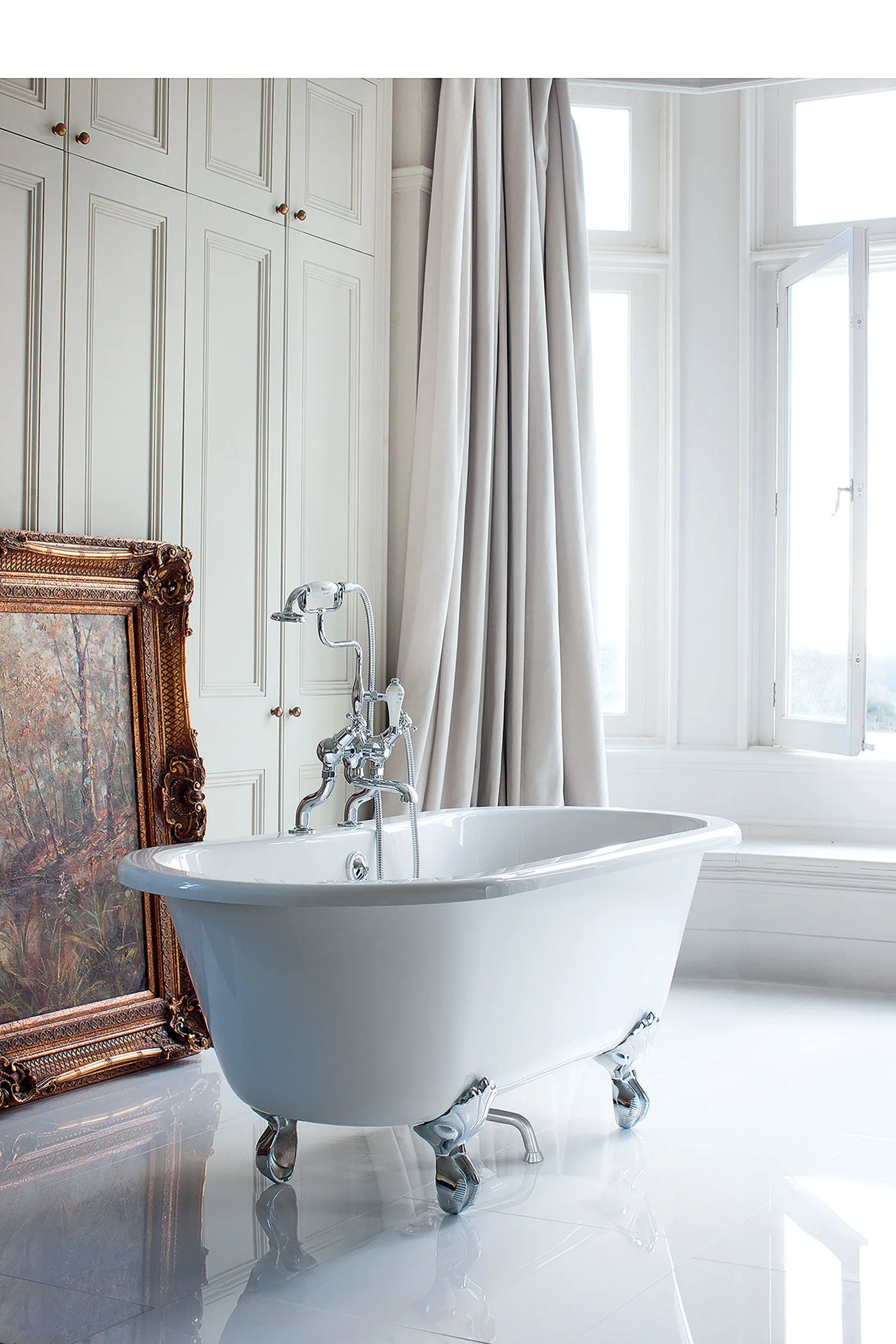 White claw-foot bath beside tall windows, pale curtains, paneled cabinets and a large leaning framed picture.