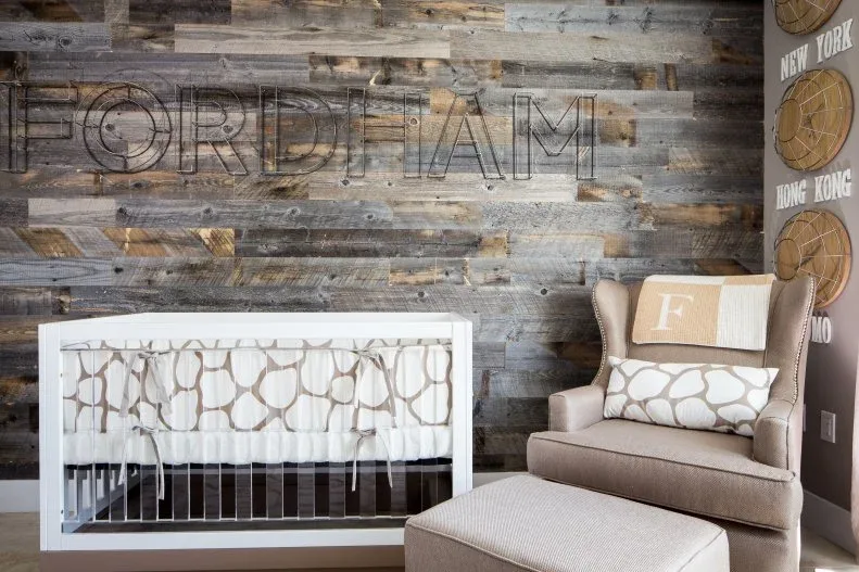 Pale crib and upholstered adult chair against varied gray-and-brown wall with large name decoration and checked cushion.