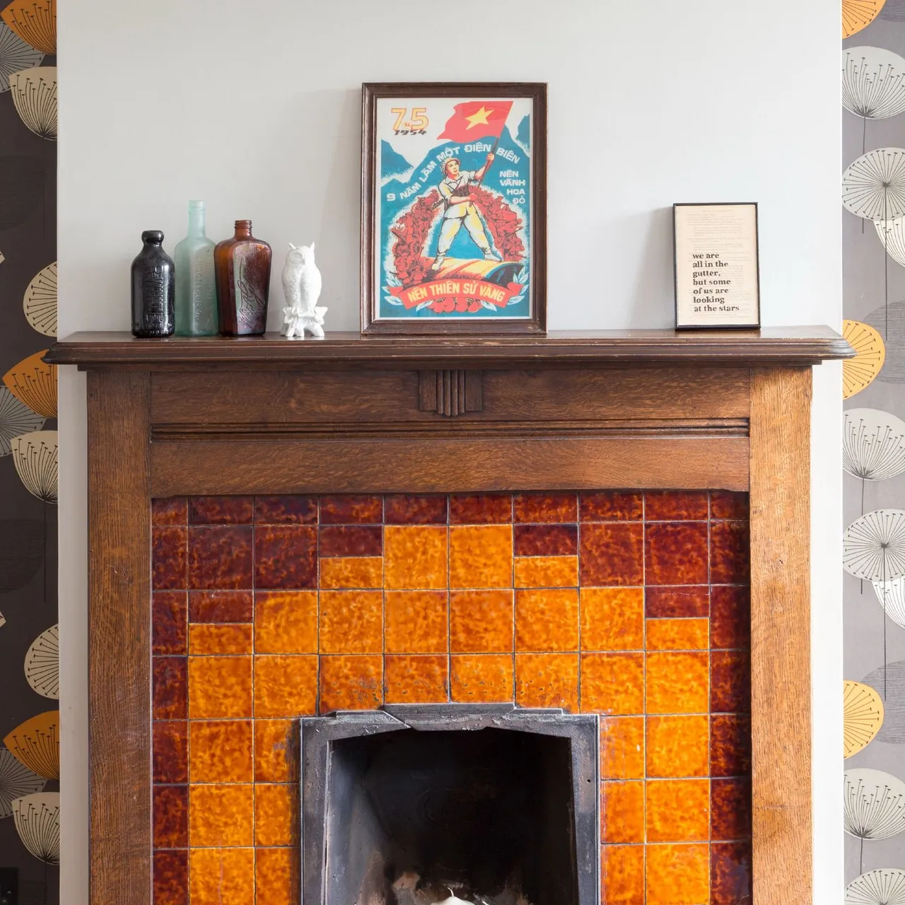 Wood mantel above orange and rust square tiles with colored bottles, white bird ornament and framed picture