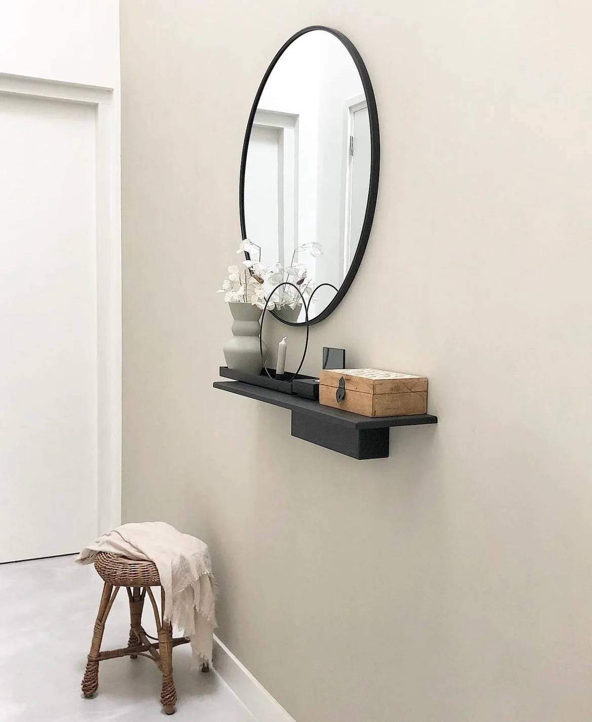 A narrow black wall shelf holds a wooden box and pale vessels beneath a round mirror beside a woven stool