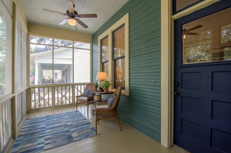 Narrow screened porch with green siding, navy door, pale trim and two woven chairs beside blue rug