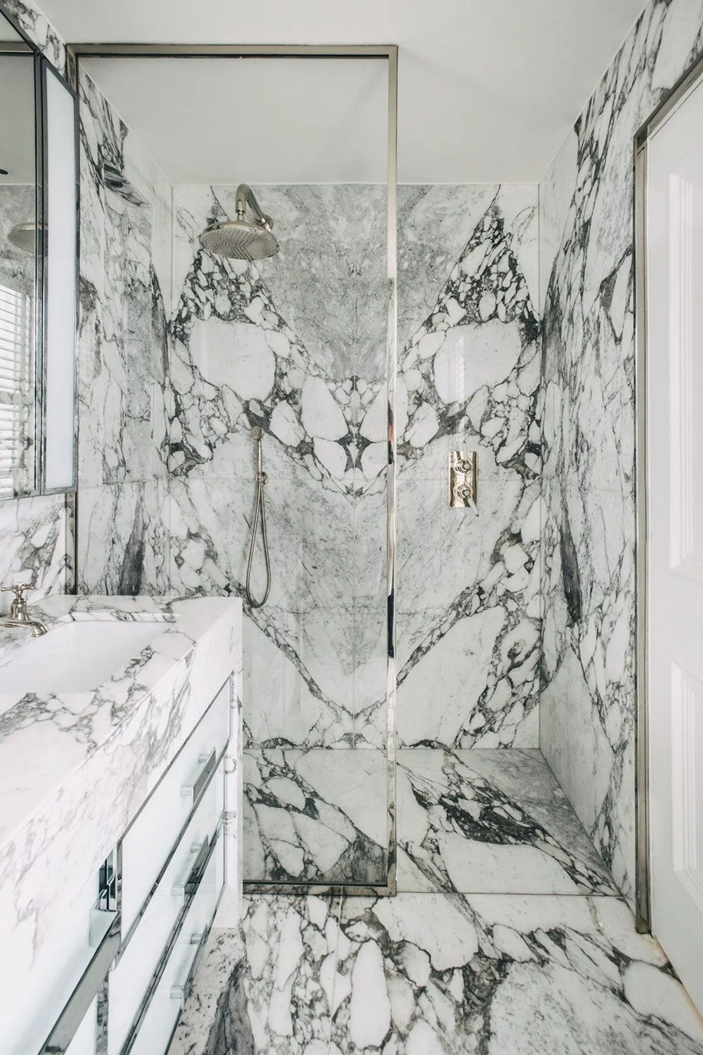 A clear shower enclosure and pale vanity counter sit within strongly veined light wall and floor surfaces beneath simple warm-toned fittings