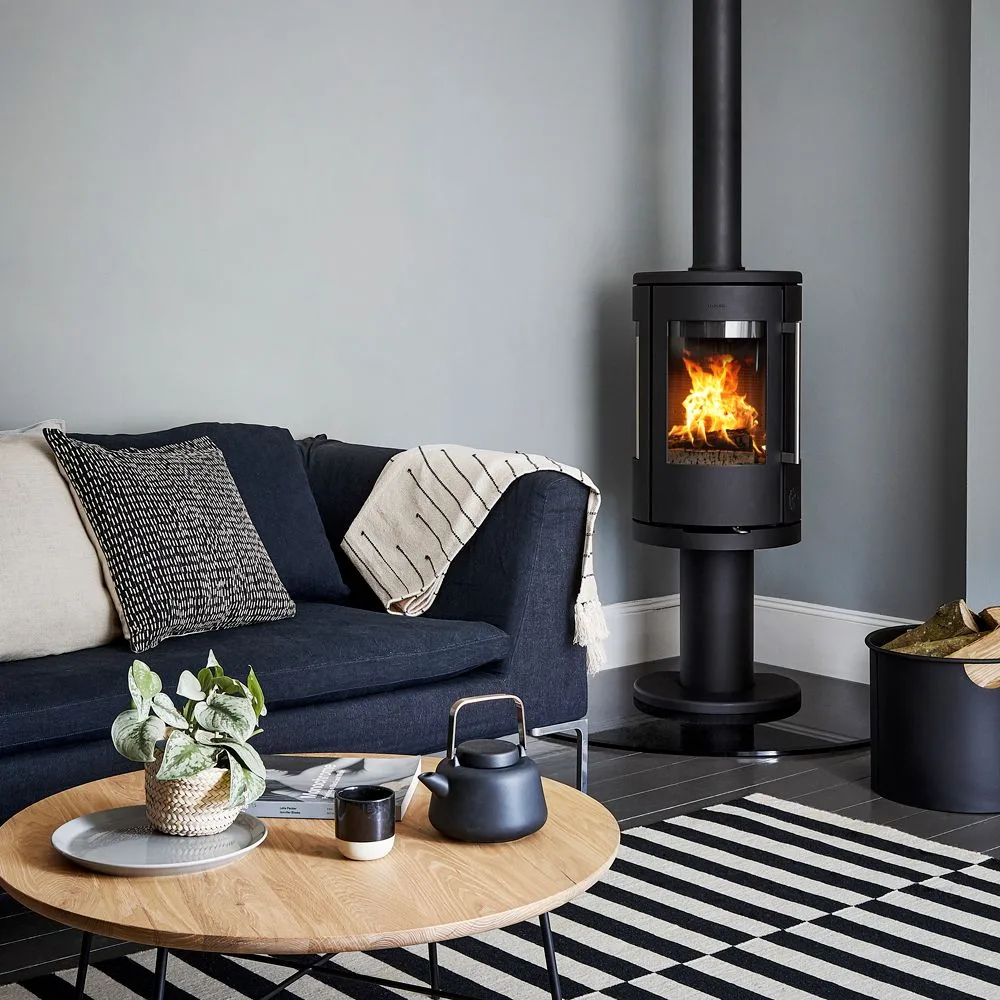 Black freestanding stove on dark platform beside blue sofa, log container and black and white rug