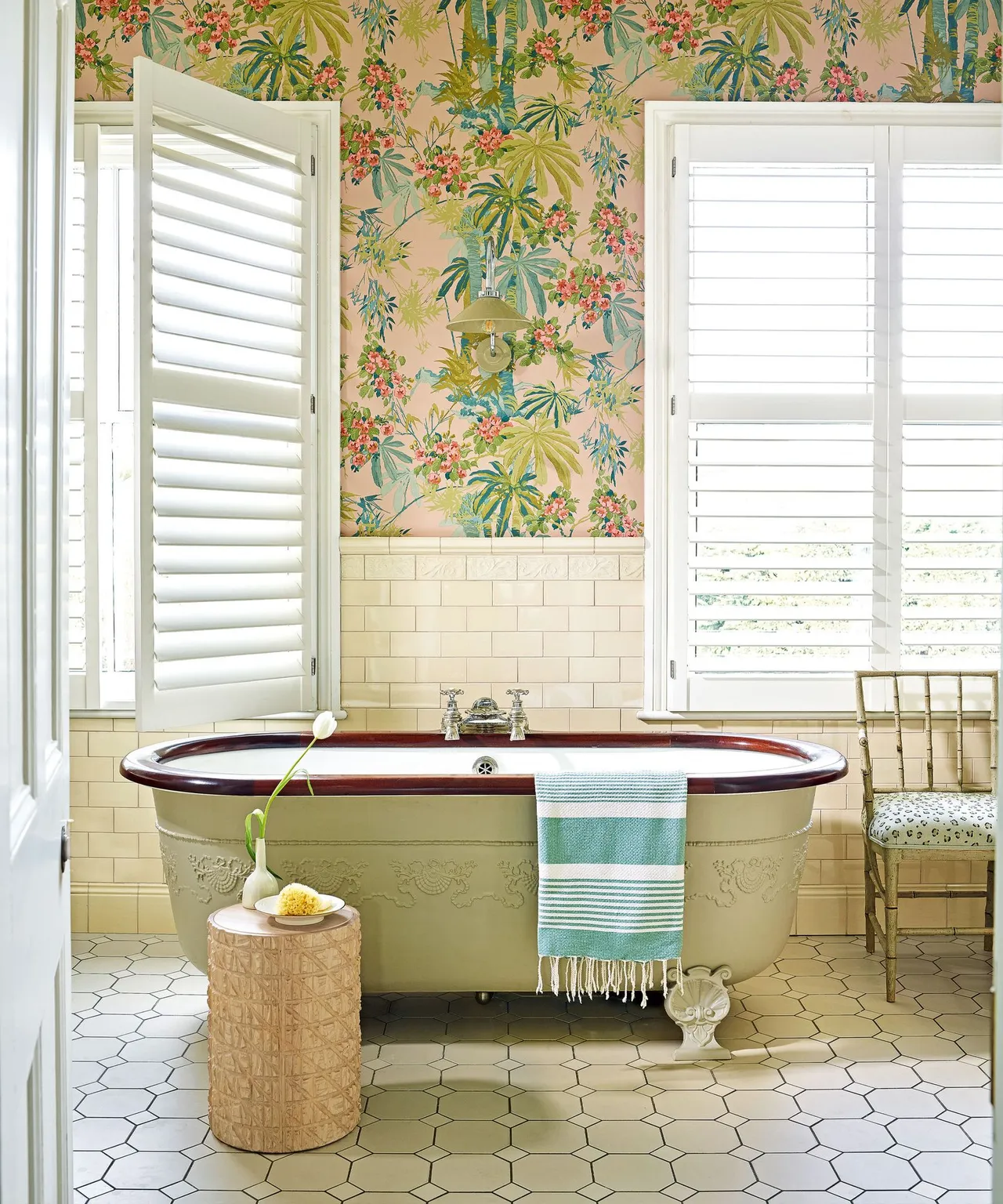 A bath sits beneath colorful leafy walls between bright windows, with a woven-looking stool nearby.